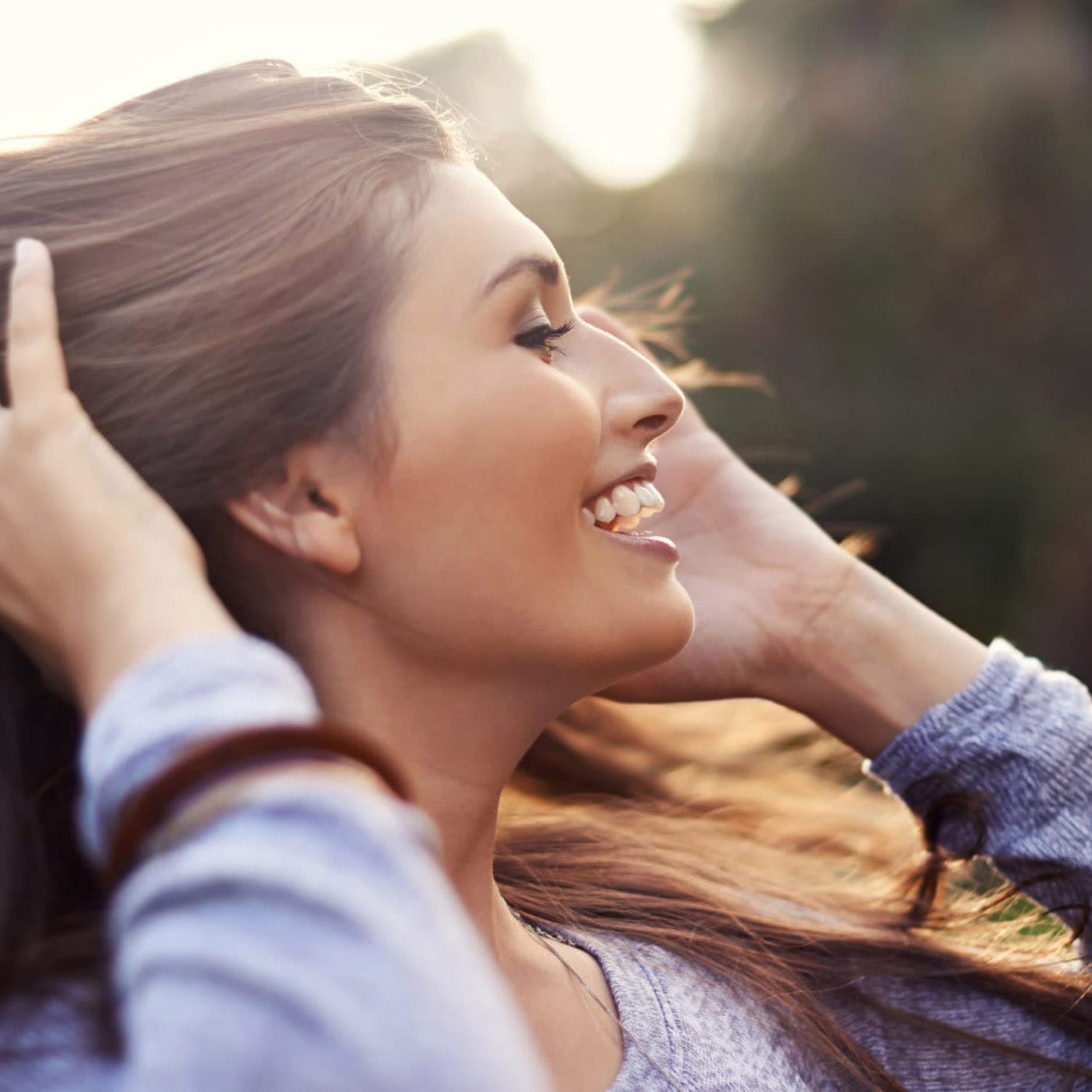smiling woman with long brown hair and blue shirt holding her hair in the wind